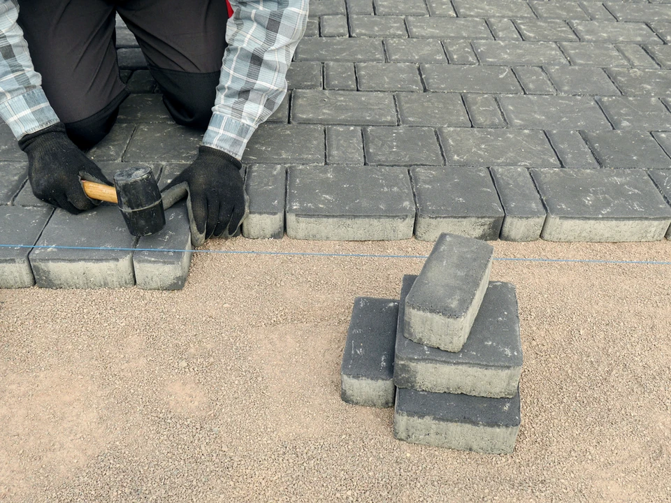 Ouvrier en tenue de travail installant un pavage extérieur, agenouillé sur surface bétonnée avec marteau et gants de protection