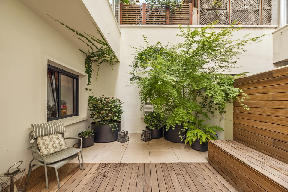 Terrasse extérieure aménagée avec plusieurs jardinières, bacs à fleurs et suspensions florales contenant des fleurs colorées et variées (roses, géraniums, pensées, piétunias, etc.). La terrasse pavée ou en bois accueille du mobilier discret, créant un espace détente verdoyant et fleuri. La composition montre une sélection soigneuse de plantes en pots de différentes tailles et hauteurs, créant une atmosphère apaisante et élégante.